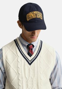 Mies, joka on pukeutunut tummansiniseen lippalakkiin, jossa on teksti "GANT 1949", vaaleanbeigeen pystykudottuun villapaitaan, jossa on tummansiniset raidat, siniraitaisiin paitaan ja punasiin sekä tummansinisiin raitoihin, katsoo alaspäin.