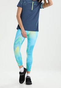 Modrý sportovní top s krátkým zipem a geometrickým vzorem, spárovaný s živě modrými legínami s abstraktními zelenými a žlutými motivy. Černé tenisky.