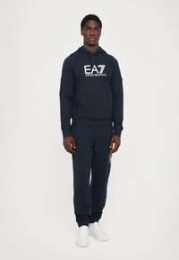 Felpa navy con tasca frontale e logo EA7 bianco, abbinata a pantaloni joggers coordinati con accentuazione del logo. Completata con sneakers bianche.