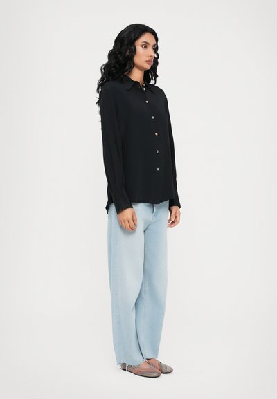 Blusa preta de mangas longas com frente de botões, combinada com jeans de perna larga azul-clara e sapatos rasteiros listados, exibindo uma silhueta descontraída.