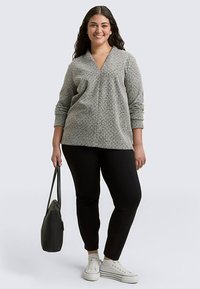 Gray textured pullover with a V-neck, long sleeves, paired with black trousers and white sneakers. Model holds a black tote bag.