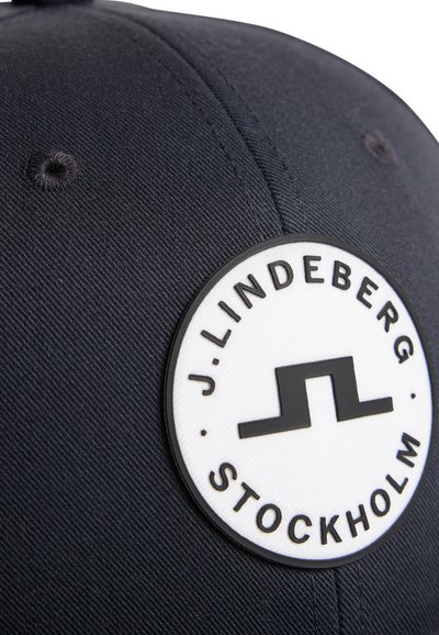 Ciemnogranatowa czapka z okrągłą białą naszywką z czarnym napisem "J. LINDEBERG STOCKHOLM" i stylizowanym czarnym logo na środku.