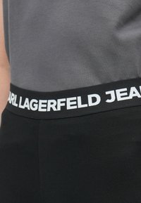 Juodos leginsai su plačiu elastingų juosmeniu, ant kurio didelėmis baltomis raidėmis parašyta "CARL LAGERFELD JEANS". Minkšta, elastinga medžiaga su glotnia tekstūra.
