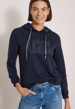 Smilende kvinde med kort brunt hår iført navy hættetrøje med snore og "CHASE THE LIGHTS" tekst, kombineret med blå jeans, hånd i lommen.
