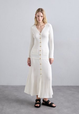 Witte gebreide cardigan met gouden knopen, gecombineerd met een bijpassende geplooide rok. De textuur heeft een patroon, aangevuld met zwarte sandalen.
