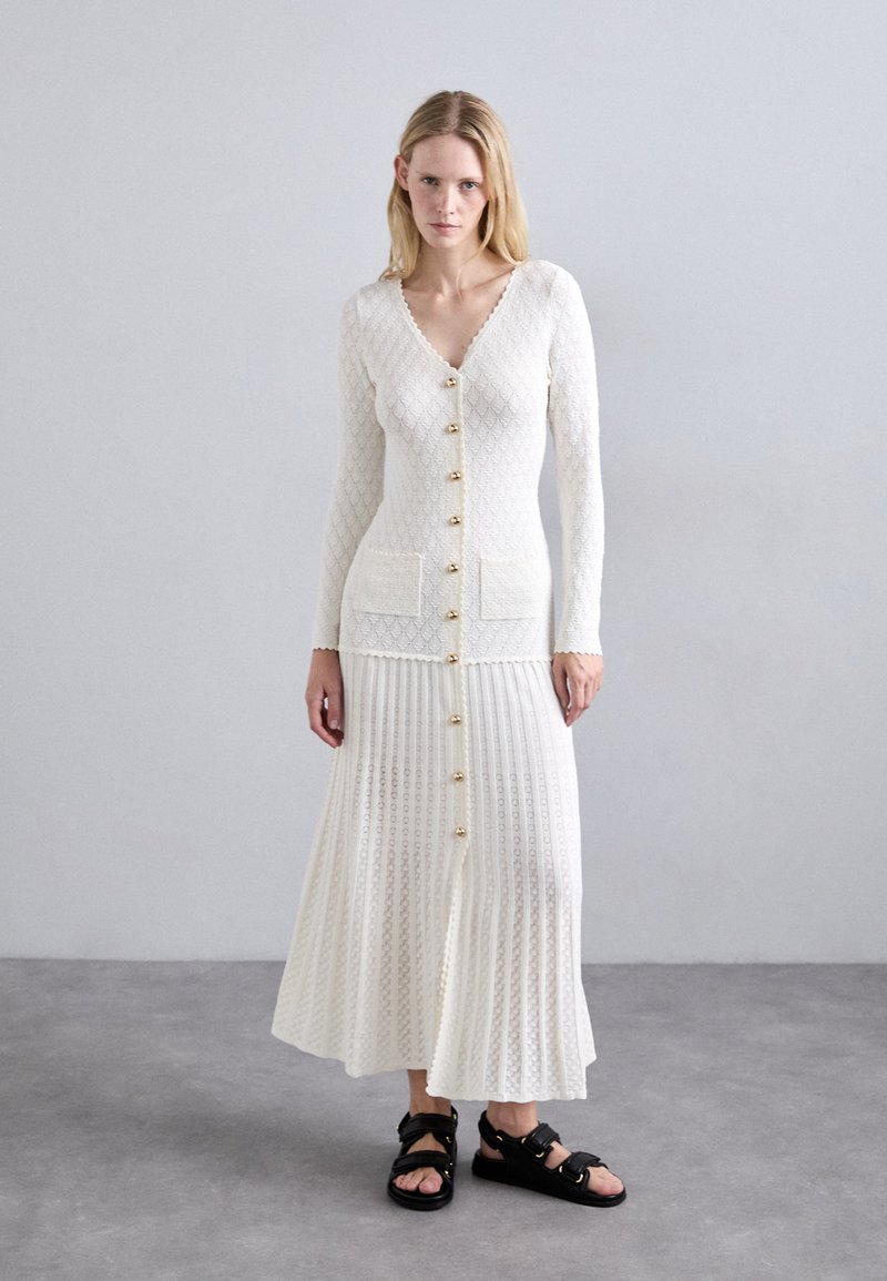 Witte gebreide cardigan met gouden knopen, gecombineerd met een bijpassende geplooide rok. De textuur heeft een patroon, aangevuld met zwarte sandalen.