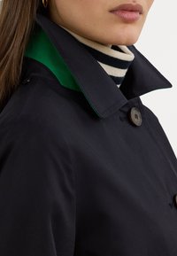 Marinblå trench coat med grön krage, med knappstängning och en randig polotröja under. Slät tyg och strukturerad design.