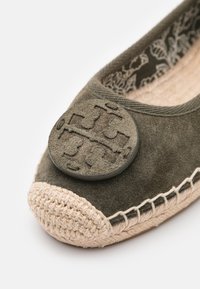 Chaussure en daim vert olive avec un emblème rond et texturé de couleur olive sur le bout. Dispose d'une semelle style jute tissée et de coutures décoratives.