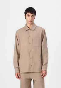 Camisa beige de manga larga hecha de tela acanalada. Presenta un cuello con solapa, una parte delantera con botones y un bolsillo de parche en el lado izquierdo.