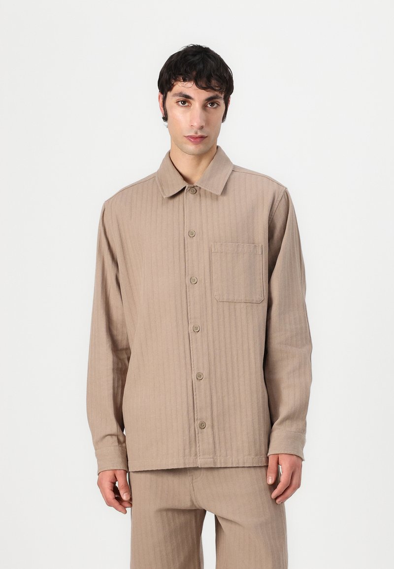 Camisa beige de manga larga hecha de tela acanalada. Presenta un cuello con solapa, una parte delantera con botones y un bolsillo de parche en el lado izquierdo.