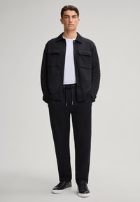 Felpa nera con bottoni sul davanti e due tasche sul petto, abbinata a pantaloni joggers neri e scarpe atletiche nere su uno sfondo neutro.