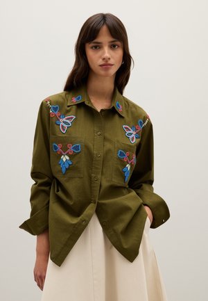 Jeune femme portant une chemise olive à boutons avec des broderies florales bleues et rouges, associée à une jupe beige, sur un fond uni.