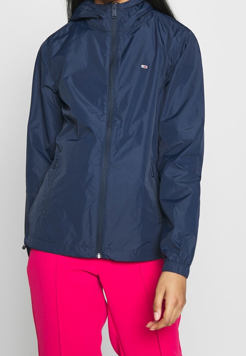 Granatowa kurtka przeciwwiatrowa z kapturem, z zamkiem z przodu, elastycznymi mankietami i bocznymi kieszeniami; lekka i gładka w dotyku. Małe logo na klatce piersiowej.
