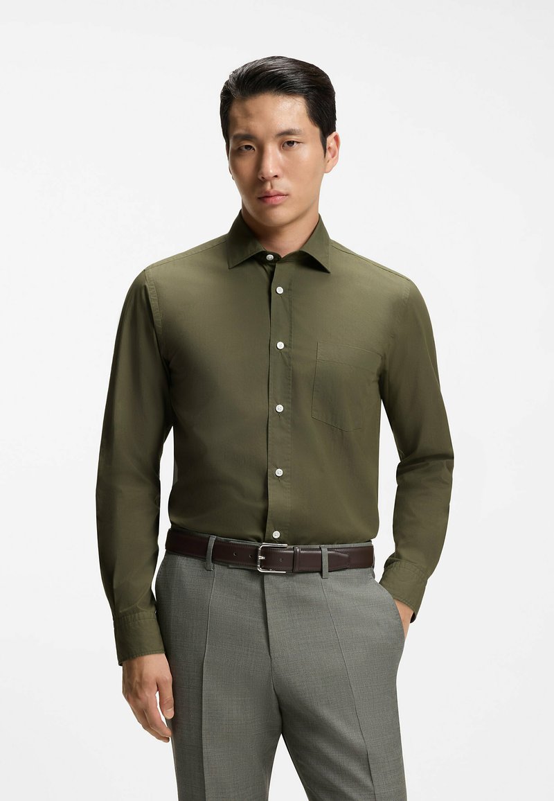 Homme portant une chemise boutonnée vert olive rentrée dans un pantalon gris avec une ceinture sombre, debout devant un fond blanc uni.