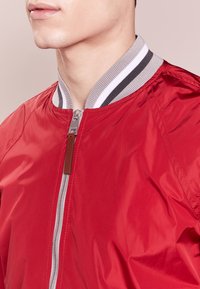 Primo piano di una persona che indossa una giacca bomber rossa con un colletto a righe grigie, bianche e nere e una zip argentata con un tirante in pelle.