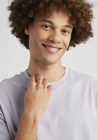 Bague en argent avec des accents décoratifs portée au poignet gauche, tenue près du cou. T-shirt gris clair, modèle avec des cheveux bouclés et un sourire subtil.