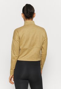 Khaki pulovr s vysokým límcem, strukturovanou tkaninou a kleslými ramenními švy, kombinovaný s černými legínami s vysokým pasem. Zobrazeno ze zadní strany.