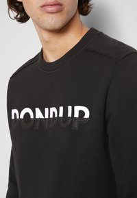 Homme portant un sweat-shirt noir avec le mot « DONDUP » imprimé en blanc et noir sur la poitrine, visage et épaules partiellement visibles.