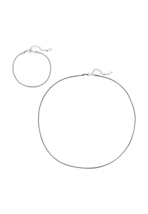 Mann trägt ein weißes, geripptes Tanktop und eine dünne silberne Halskette, zeigt Hals- und oberen Brustbereich.