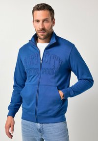Sudadera azul con cremallera y bordado "PORT CITY LIVERPOOL" en un tono azul más oscuro. Cuenta con puños acanalados y bolsillos, confeccionada con una tela suave.