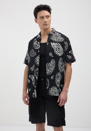 Homme portant une chemise à manches courtes noire et blanche à motif paisley sur un débardeur noir et un short cargo noir, regardant sur le côté contre un fond uni.