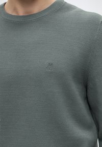 Gros plan d’une personne portant un pull vert texturé à col rond avec un petit logo brodé sur la poitrine gauche.