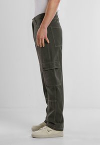 Pantalones cargo de color verde oliva oscuro con múltiples bolsillos, detalles de costura visibles, ajuste entallado y una textura suave, combinados con zapatillas de deporte color blanco roto.