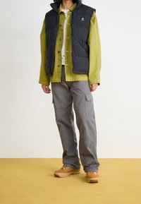 Czarna, puchowa kamizelka, zielona koszula, szare spodnie bojówki. Beżowe buty z żółtymi akcentami i sznurówkami. Prosty design z zapięciem na guziki i detalami kieszeni.