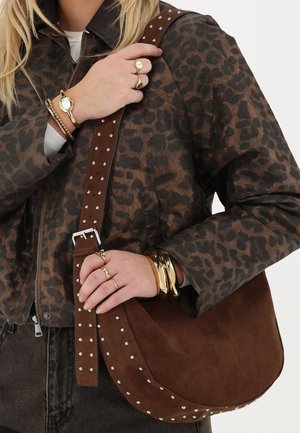 Bruine suède schoudertas met zilveren studs langs de band, gedragen bij een jas met luipaardprint. Gouden sieraden aan de hand.