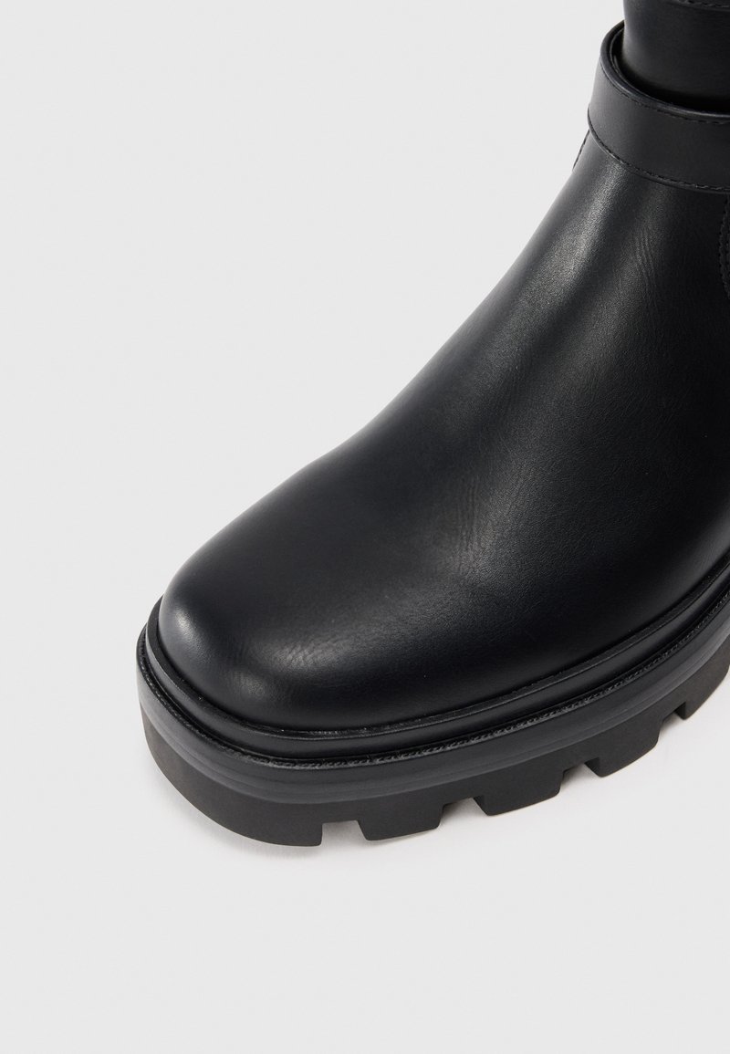Botín de cuero negro hasta el tobillo con punta redondeada, suela de goma negra gruesa y textura suave. Presenta un detalle de correa sencilla en el tobillo.