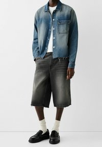 Giubbotto di jeans in blu chiaro sbiadito con bottoni e dettagli nelle tasche; abbinato a pantaloni corti in denim neri e mocassini neri. Calzini bianchi a vista.