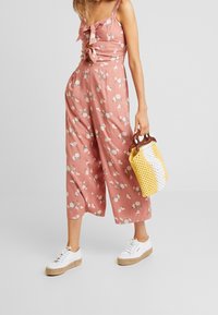 Frau trägt einen weiten, pinken Jumpsuit mit Blumenmuster und Bindedetails, weiße Plattform-Sneaker und hält eine gelb-weiße Strick-Handtasche.