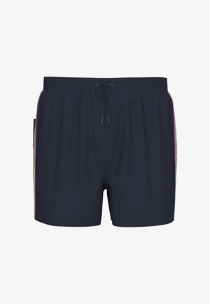 Jeune homme debout pieds nus, portant un short de bain bleu marine avec des bandes latérales blanches et rouges, sur un fond gris uni.