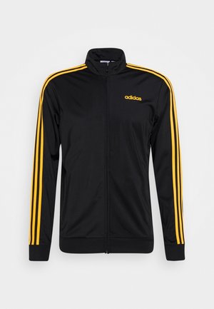 Czarna kurtka sportowa Adidas z żółtymi paskami na rękawach i żółtym logo Adidas na lewej piersi, zapinana z przodu na zamek błyskawiczny, z kołnierzem typu stójka.