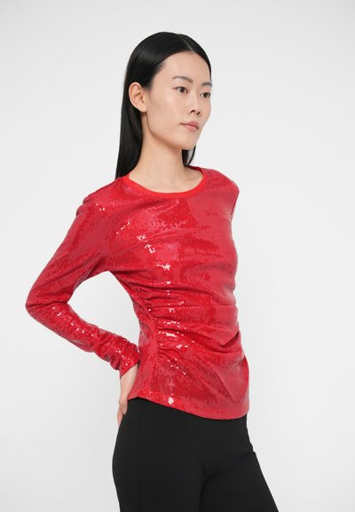 Punane pikkade varrukatega särk, mis on kaetud litritega ja millel on ümmargune kaelus. Särk on istuv ning selle pind on sile. Alt on näha mustad püksid.