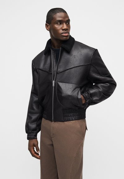 Άνδρας που κοιτάζει προς τα αριστερά, φοράει μαύρο δερμάτινο μπουφάν με φερμουάρ και γιακά από shearling, με καφέ παντελόνι, το ένα χέρι στην τσέπη του μπουφάν.