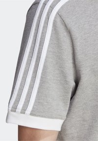 Graues Sportshirt mit weißem Drei-Streifen-Design an den Ärmeln, geripptem weißen Kragen und strukturiertem Stoff.