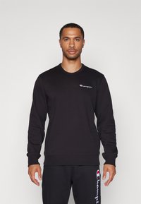 Sweatshirt noir à manches longues, col rond, avec des poignets et un ourlet côtelés. Il possède un petit logo brodé sur la poitrine et une texture lisse.