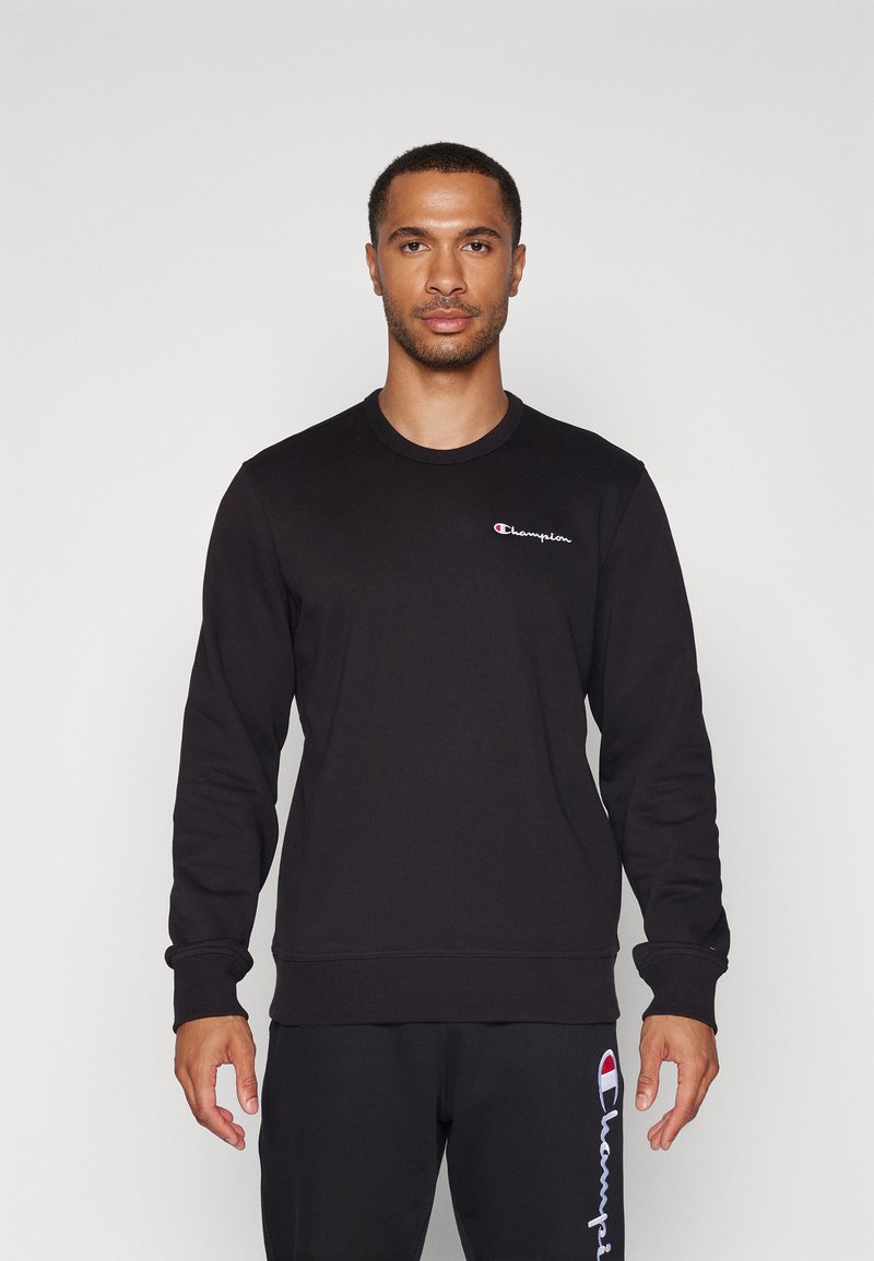 Sweatshirt noir à manches longues, col rond, avec des poignets et un ourlet côtelés. Il possède un petit logo brodé sur la poitrine et une texture lisse.
