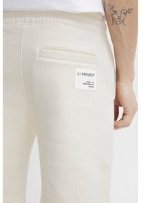 Beige treningsbukser med en myk tekstur, elastisk midjebånd og baklomme med en hvit etikett som har "11 PROJECT"-merket.