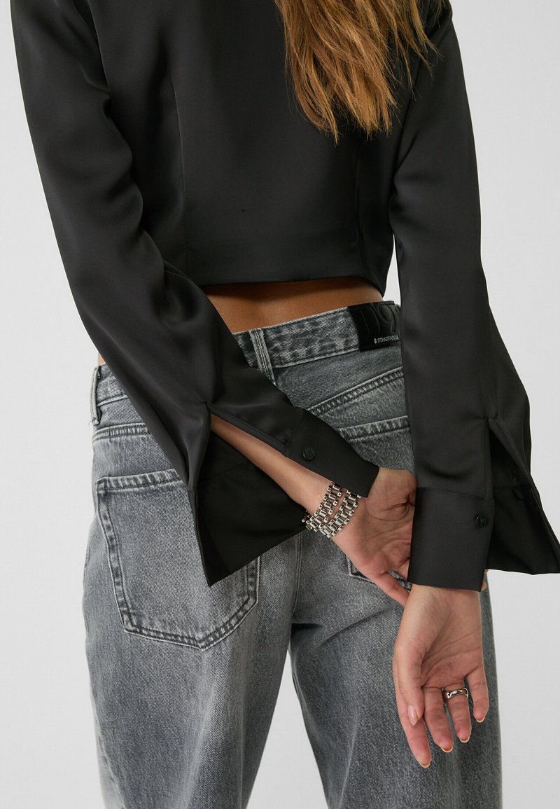 Blouse courte en satin noir à manches larges, associée à un jean en denim gris délavé taille haute. Bracelet argenté au poignet.