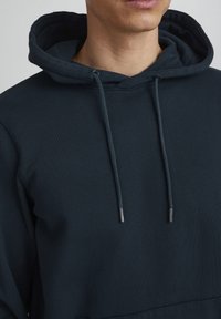 Tumši zils kapucis ar teksturētu apdari, auklām ar metāla galiem un kengura kabatu. Audums izskatās mīksts un izturīgs.