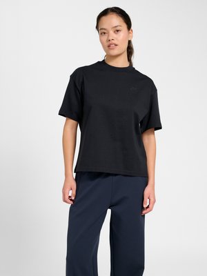 Jeune femme aux cheveux attachés, portant un t-shirt noir à manches courtes et un pantalon bleu foncé, debout devant un fond clair uni.