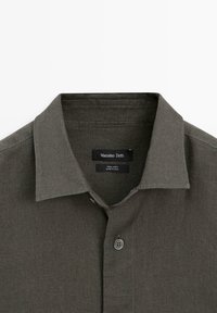 Mørkegrøn skjortekrave i hør med øverste knap lukket, der viser etiket med "Massimo Dutti" og mærkat med "100% hør, lavet i Kina" indeni.