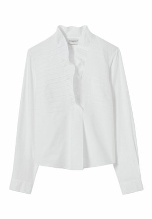 Jeune femme aux cheveux raides et foncés porte un chemisier blanc plissé, un jean bleu, des bottines marron, debout devant un fond uni gris clair.