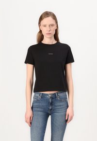 Tricou scurt de culoare neagră, din bumbac, cu un decolteu rotund și mâneci scurte. Logo imprimat în alb pe piept. Asortat cu blugi albaștri.