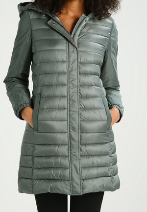 Kvinde iført knælængde, quiltet olivengrøn dunjakke med hætte, frontlynlås og hænderne i lommerne.