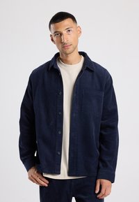 Navy corduroy jacket featuring a button-up front, spread collar, and patch pocket. Soft texture with vertical ribbing, worn over a light shirt.