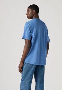 Tricou albastru din bumbac cu guler rotund și mâneci scurte, asortat cu blugi deschiși la culoare. Materialul pare neted și fără ornamente.