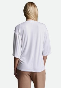 Weißes Kurzarmhemd mit entspanntem Schnitt, glatter Textur und vertikaler Nahtdetails auf der Rückseite, kombiniert mit beigen strukturierten Hosen.